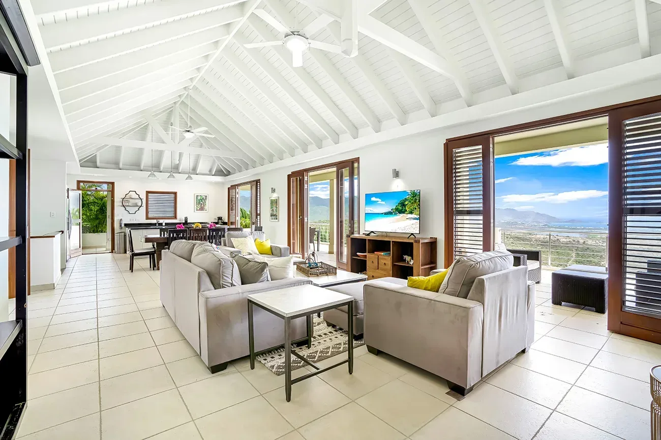 Spacious living room with gray sofas, yellow accent pillows, white vaulted ceiling, and panoramic ocean-mountain views