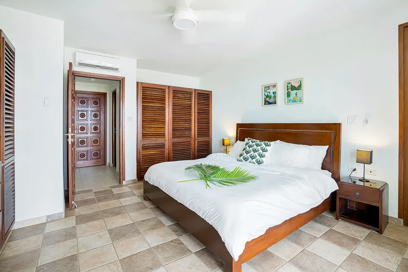 Master bedroom with wooden bed, white linens, tropical artwork, palm leaf decor, and carved wood doors