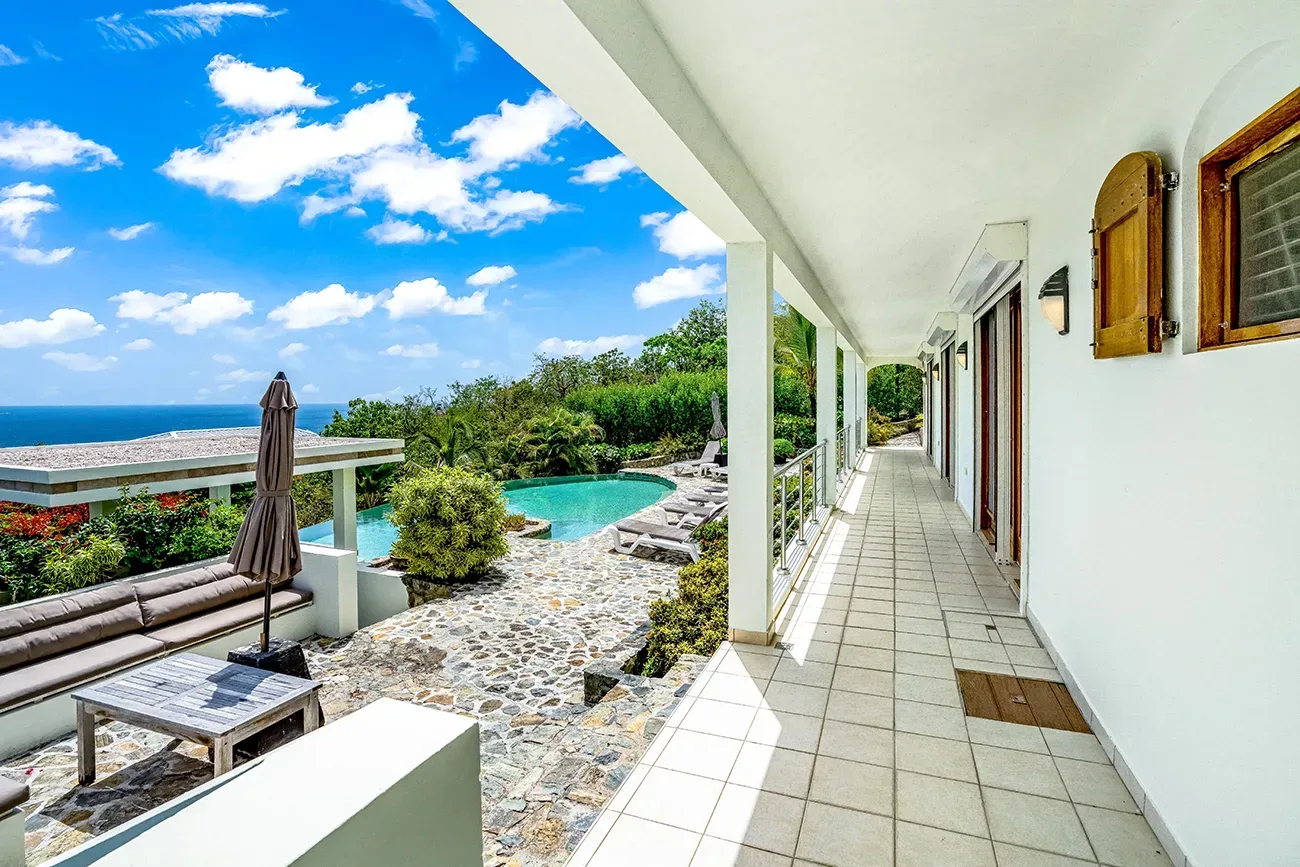 Villa corridor with ocean view, pool, stone patio, and lounge furniture under blue sky