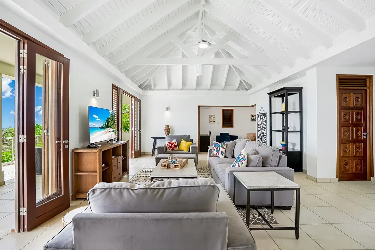 Living room with gray sofas, wooden TV stand, French doors leading to balcony with ocean view and colorful cushions