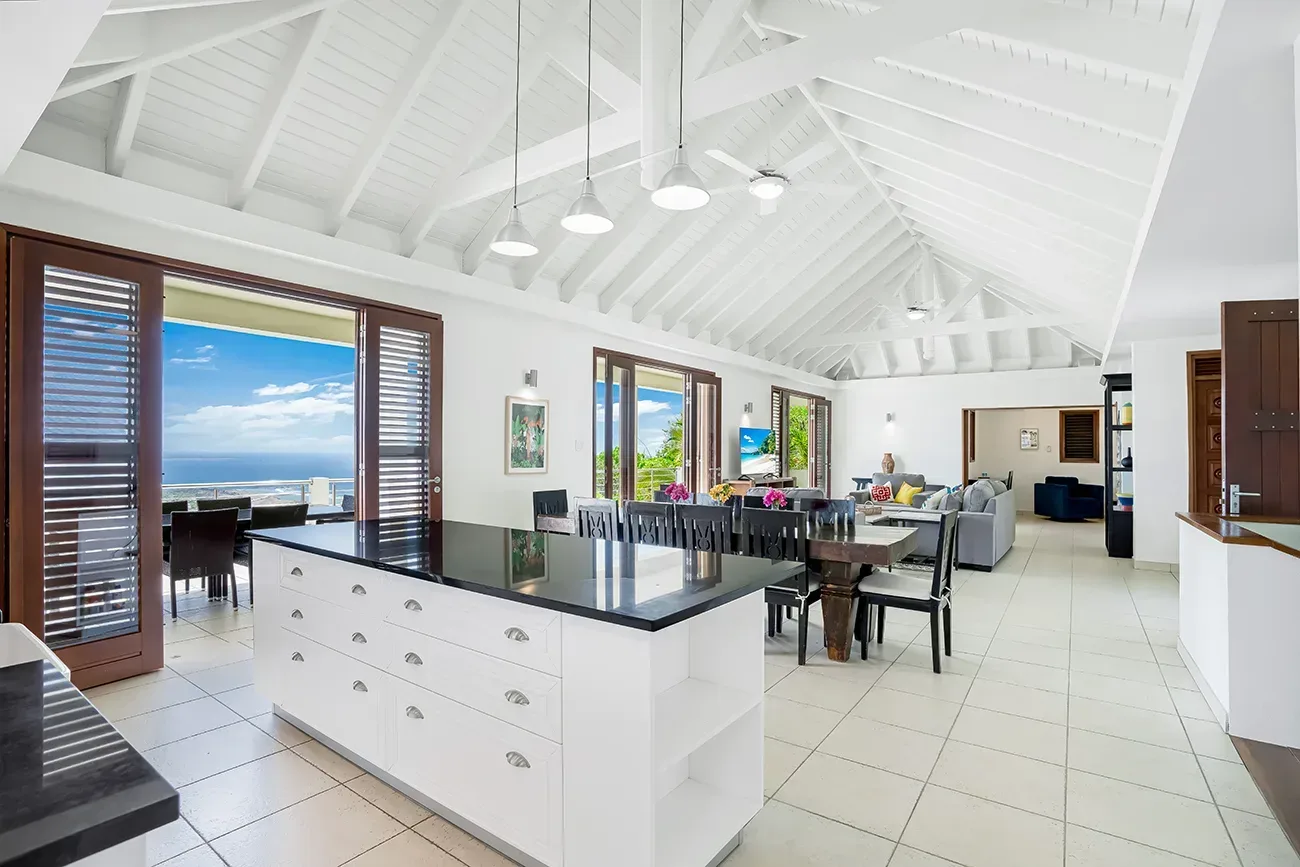 Open-concept villa interior with kitchen island, dining area, living room, and ocean view through open doors