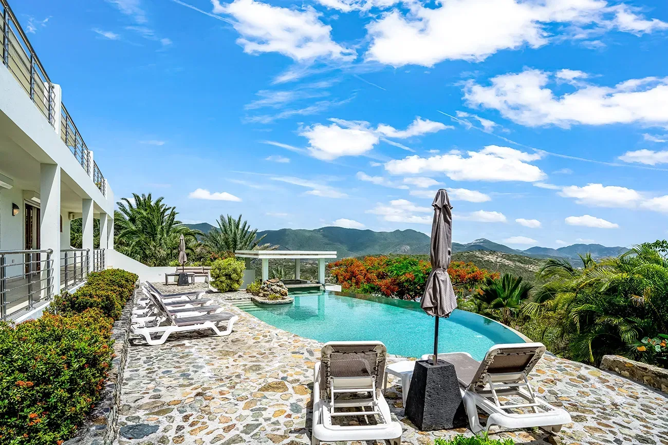 Luxury villa with turquoise pool, white lounge chairs, stone patio, parasol, and mountain views