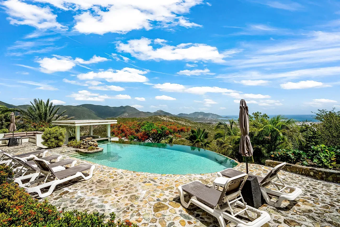 Villa terrace with infinity pool, white lounge chairs, tropical garden, and ocean views under bright blue sky