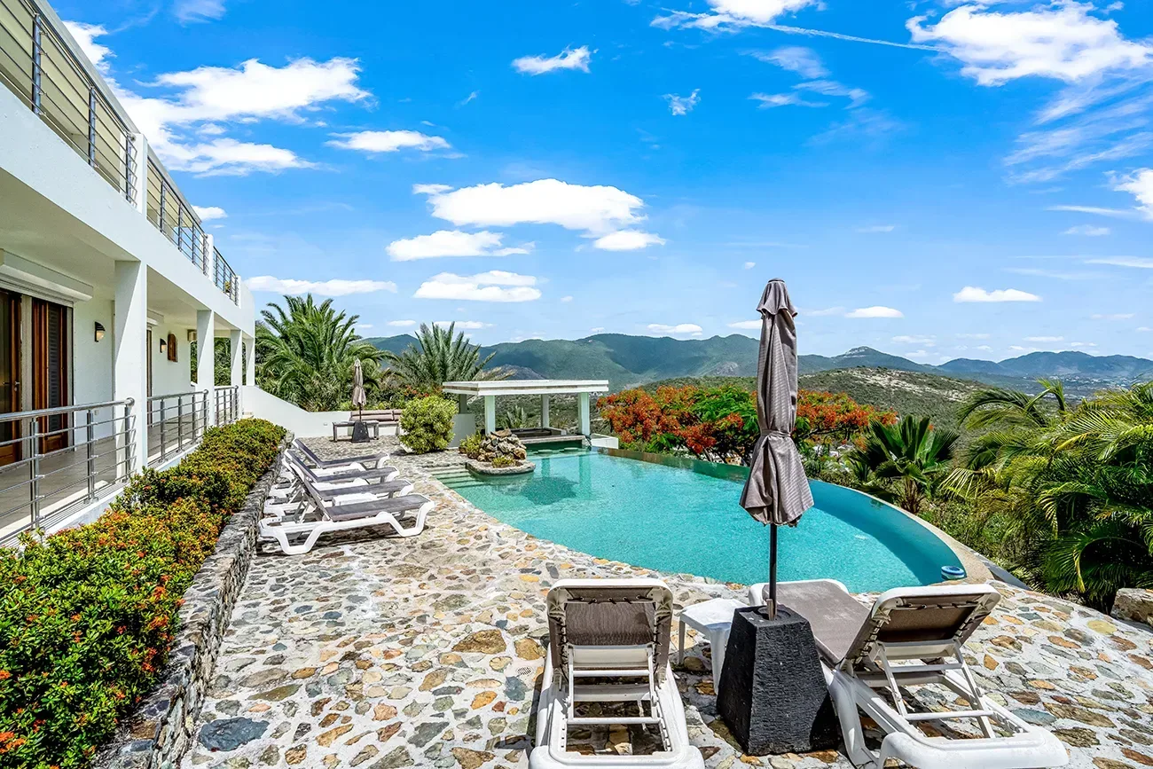 Two-story villa with turquoise pool, white lounge chairs, stone patio, and mountain views under blue sky