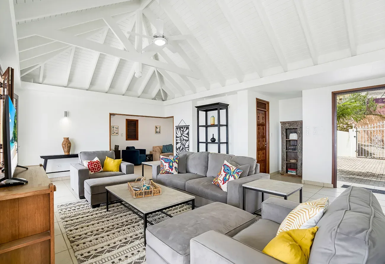 Spacious living room with gray sofas, colorful cushions, white vaulted ceiling, and outdoor view through open doors