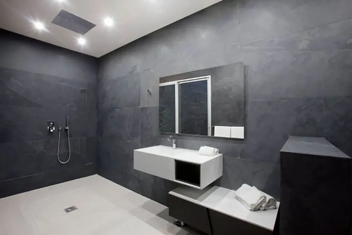 A modern minimalist bathroom with dark charcoal textured wall tiles, sleek white and grey floating vanity, and walk-in shower area.