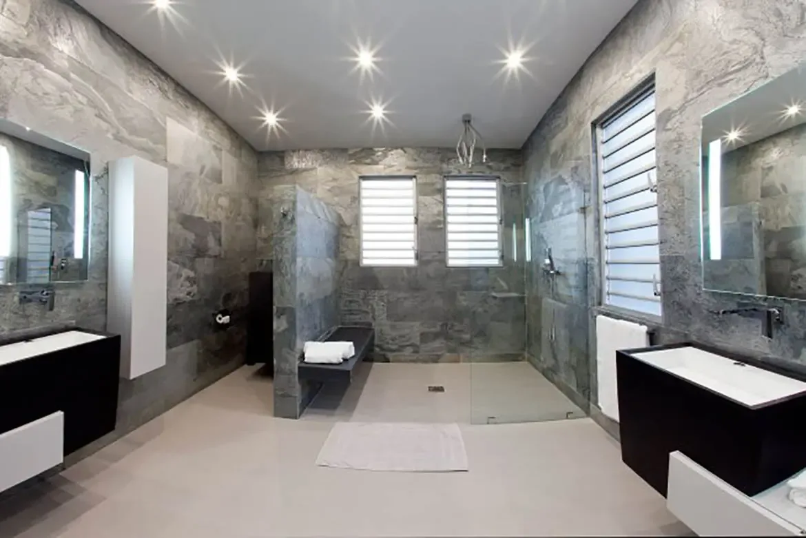 The spacious modern bathroom features grey stone-tiled walls, dual black vanities with integrated white sinks, large open shower area with glass partitions, and sleek minimalist lighting.