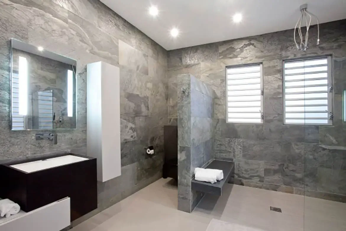 A modern minimalist bathroom showcases textured grey stone walls, dark floating vanity with white vessel sink, large backlit mirror, and walk-in shower with built-in stone bench.