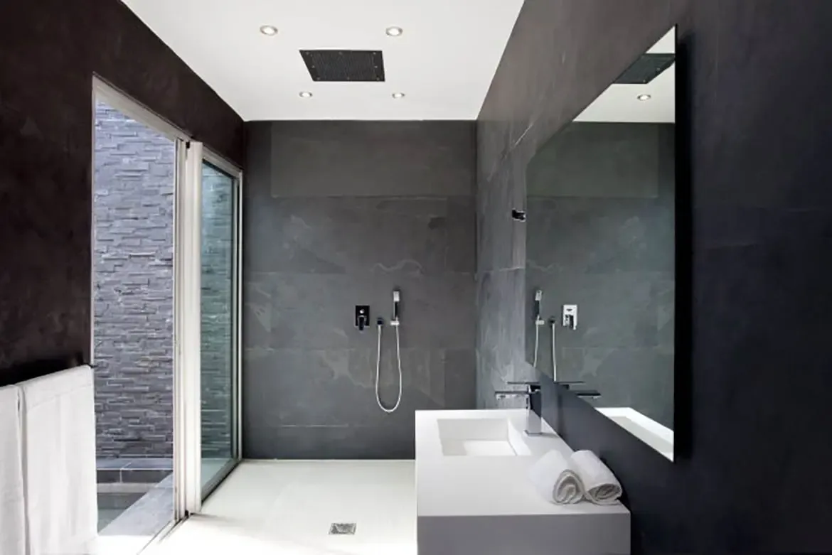 A minimalist bathroom with dark grey slate wall tiles, white integrated sink vanity, large frameless mirror, walk-in shower area, and glass door leading to outdoor space.