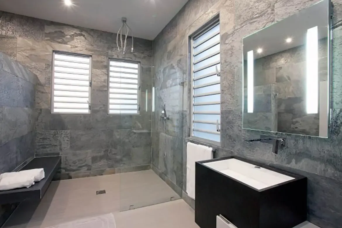 The modern bathroom features grey textured stone walls, large walk-in glass shower with bench, sleek black vanity with white rectangular sink, and large backlit mirror.