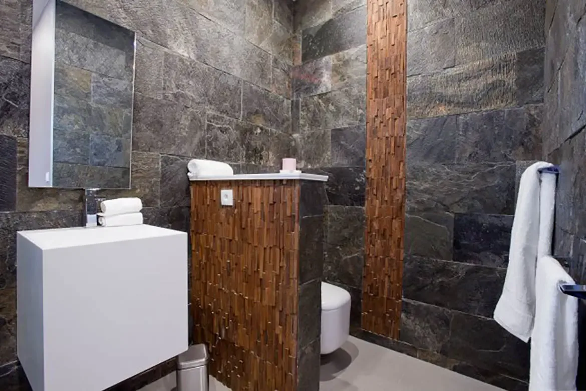 The modern bathroom showcases dark natural stone tile walls with vertical strip accent, partition wall with textured wood slats, sleek white pedestal sink, floating vanity mirror, and wall-mounted toilet.
