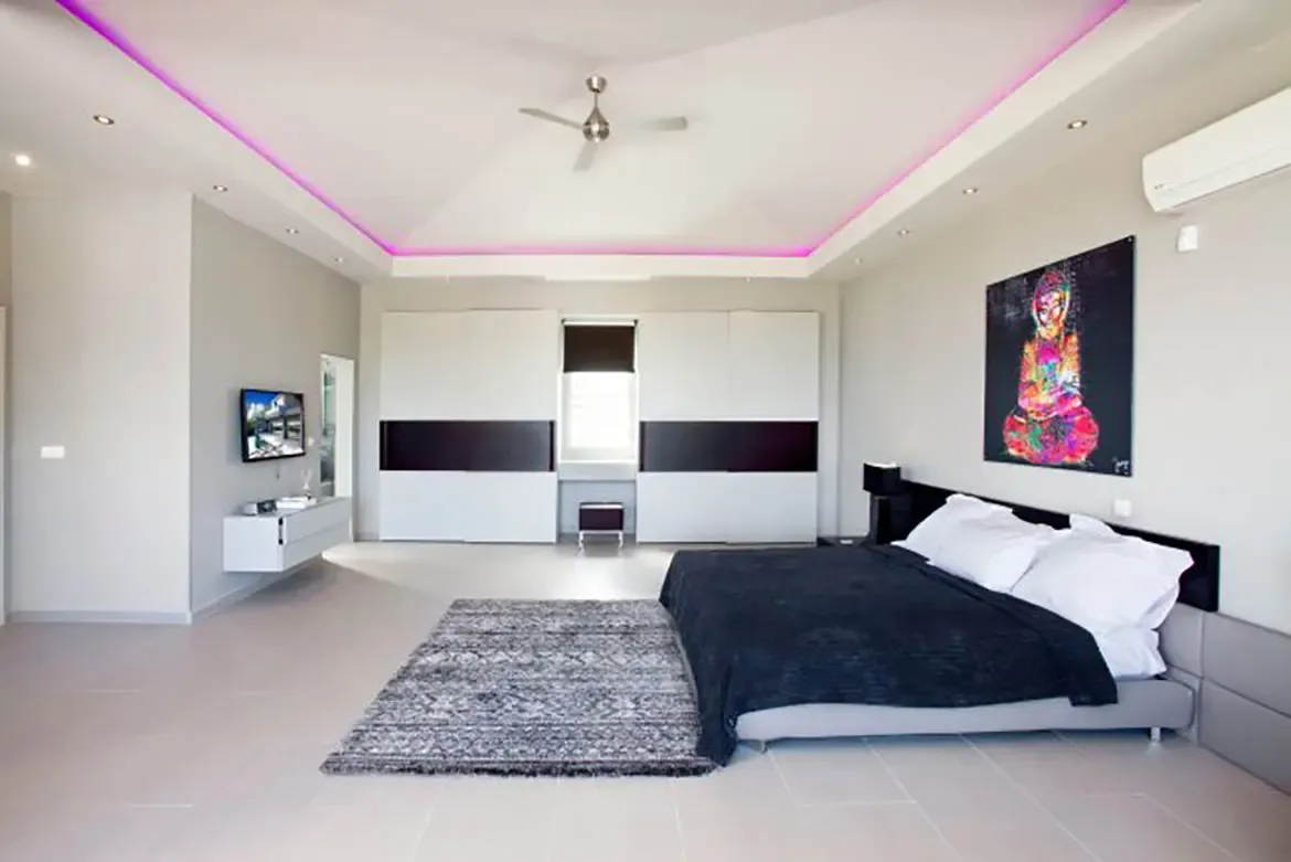 This spacious modern bedroom features a king-size bed with black bedding, grey shag rug, large colorful Buddha artwork, minimalist white built-in wardrobes, and tray ceiling with pink LED accent lighting.