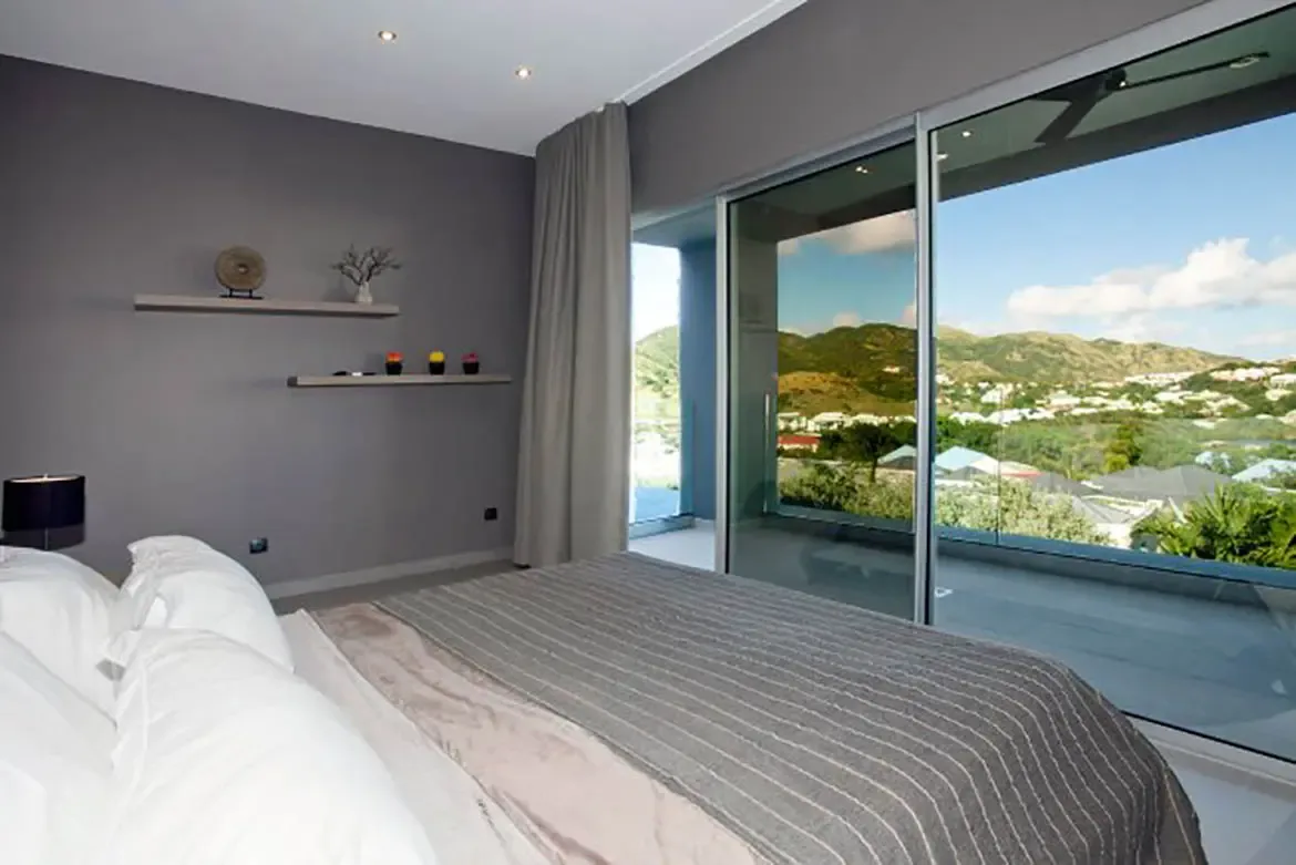 A modern bedroom features large sliding glass door opening to balcony with expansive view of rolling hills and residential valley, dark grey walls, and minimalist wall-mounted shelves.