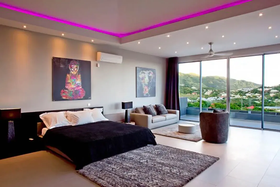The modern master suite showcases a large bed, seating area, vibrant pop-art, floor-to-ceiling windows offering panoramic mountain view, and recessed pink LED accent lighting.