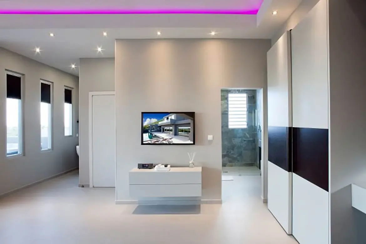 A modern minimalist bedroom with wall-mounted TV, sleek white floating console, large built-in wardrobe, pink LED accent lighting, and view into ensuite stone-tiled bathroom.