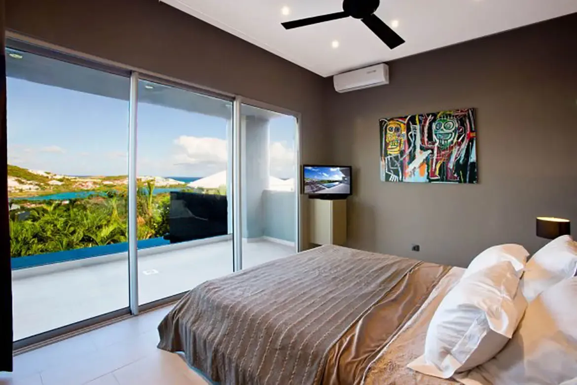 A modern bedroom with large bed, textured cover, stunning view of ocean and lush greenery through floor-to-ceiling sliding glass doors, colorful modern art on wall, and ceiling fan.