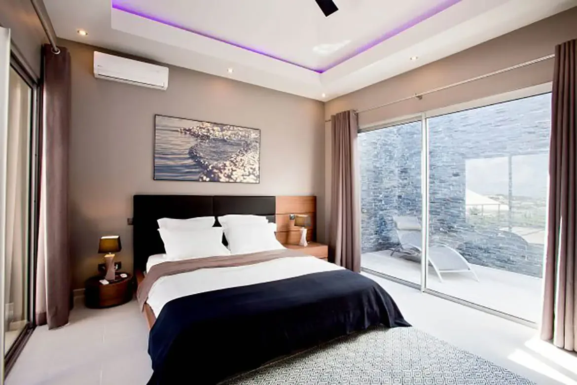 A modern master bedroom with large bed, dark headboard, crisp white linens, navy blue throw, matching bedside tables with sleek lamps, large artwork above bed, and recessed ceiling with vibrant purple LED lighting opening onto private patio.