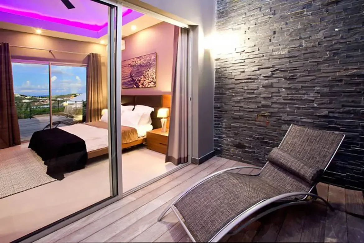A modern bedroom features purple ambient cove lighting, dark wood headboard, and large sliding glass door opening onto private terrace with textured stone accent wall and contemporary outdoor lounge chair.