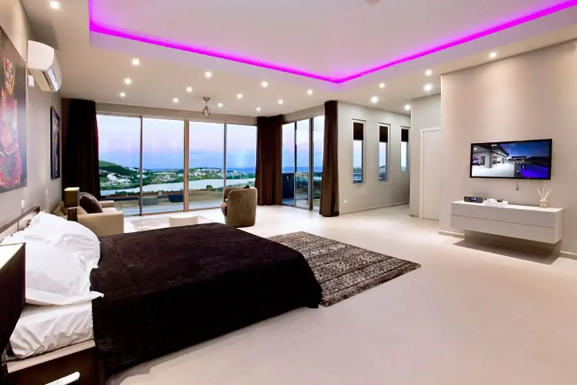 A spacious modern bedroom with floor-to-ceiling windows overlooking scenic water view, large bed, minimalist furniture, and tray ceiling with vibrant pink neon accent lighting.