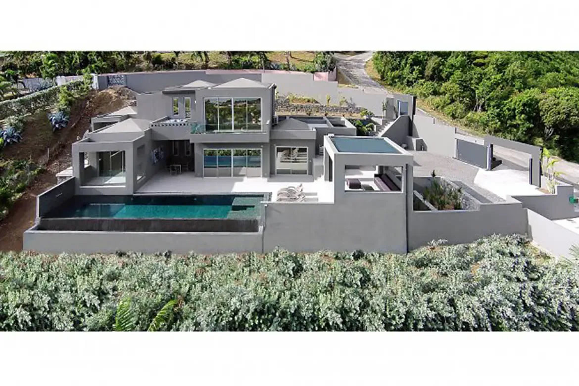 An aerial view reveals the modern multi-level grey villa with lap pool, expansive patio, and geometric architectural wings set against a lush hillside.