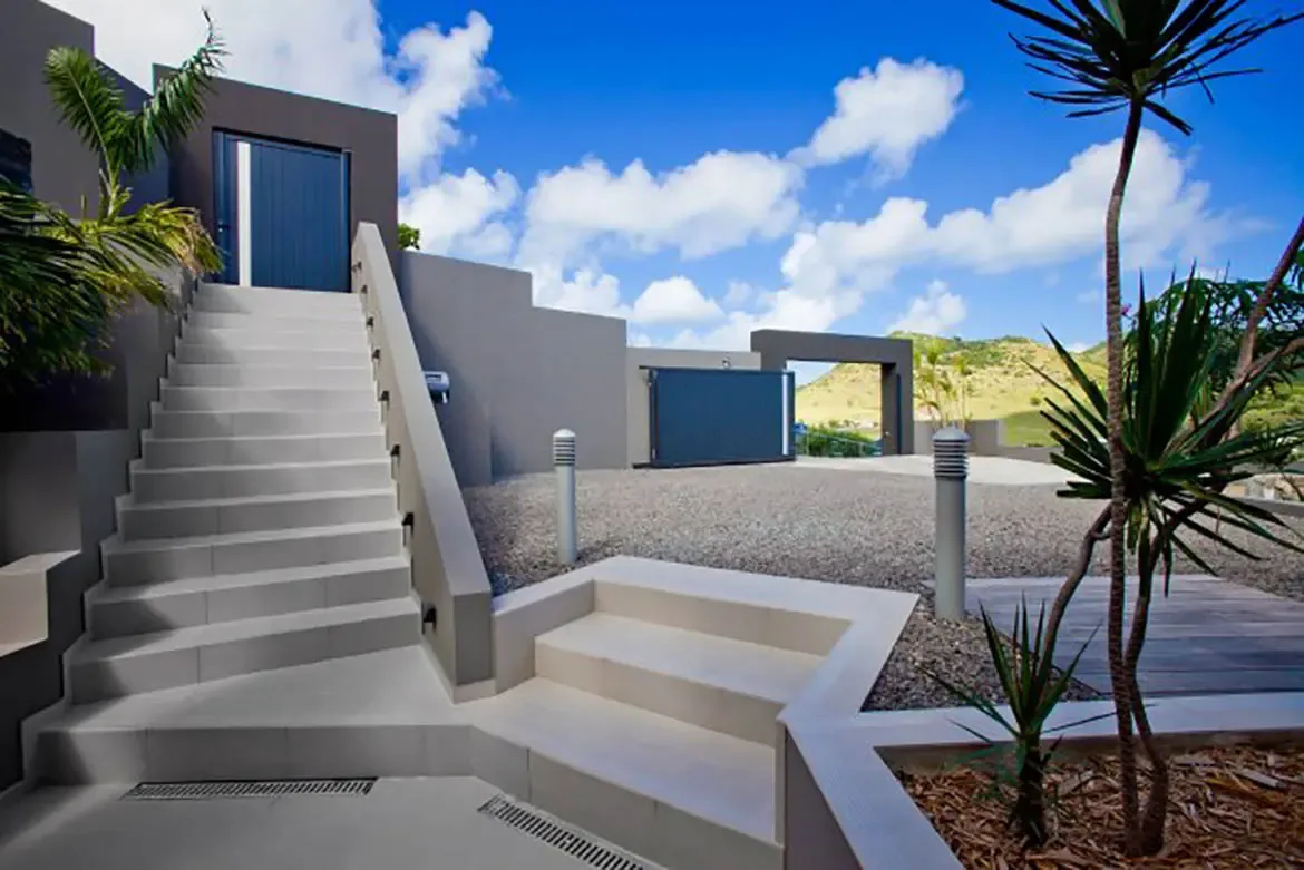 The modern villa entrance features clean-lined gray walls, light-colored stairs, gravel courtyard, tropical landscaping, and clear blue sky with distant hills view.