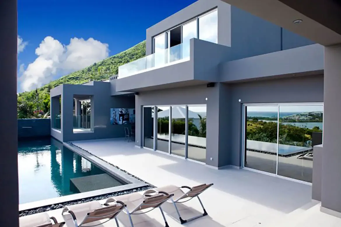 A modern gray villa showcases a large swimming pool, expansive white terrace with lounge chairs, and floor-to-ceiling glass doors revealing mountain and coastal views.