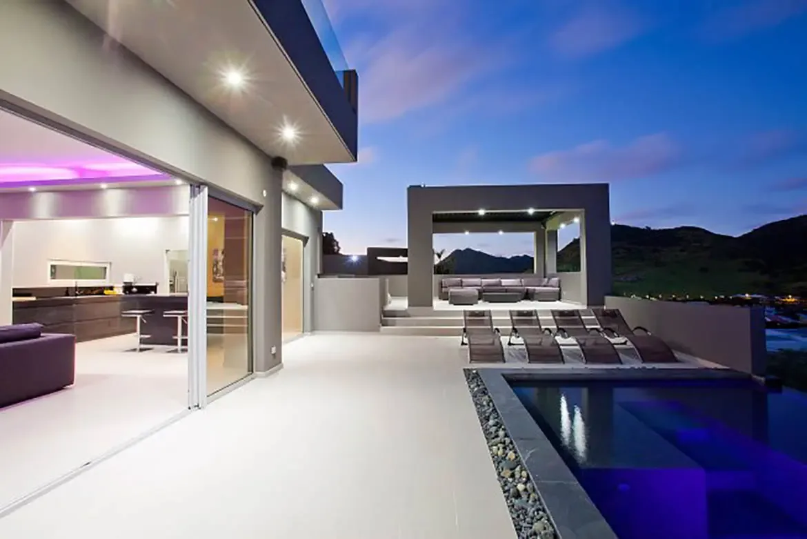A luxurious villa with seamless indoor-outdoor living, contemporary kitchen opening onto white-tiled terrace, minimalist infinity pool with purple underwater lighting, sun loungers, and covered lounge pavilion with scenic mountain views at twilight.