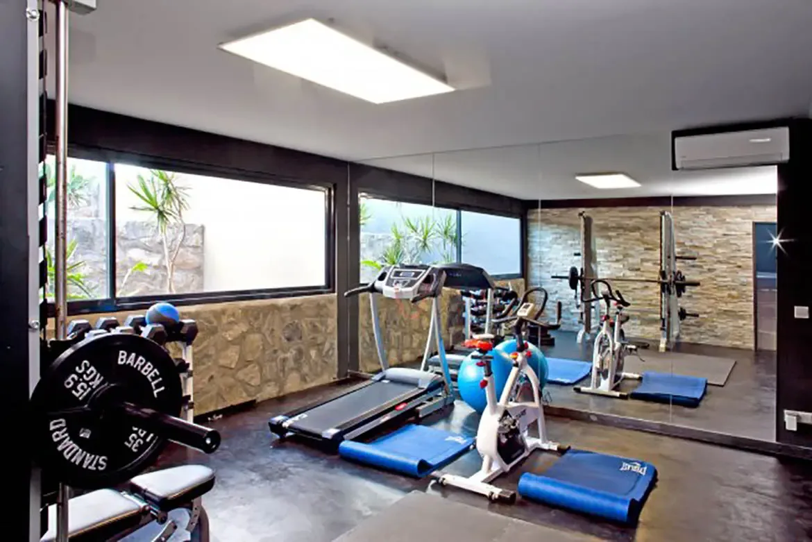 The home gym includes treadmill, stationary bike, weight bench with barbell, blue yoga mats, floor-to-ceiling mirrors, stone accent walls, and large window overlooking small garden area.