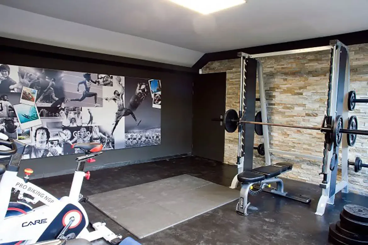 A home gym with Smith machine and weight bench, stationary bike in foreground, large wall mural depicting athletes, specialized gym flooring, and textured stone accent wall.