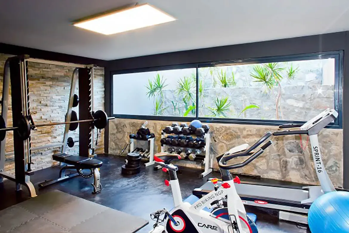 A private indoor fitness center features squat rack with bench, dumbbell rack, stationary bike, treadmill, blue exercise ball, and large windows overlooking stone-walled garden area.