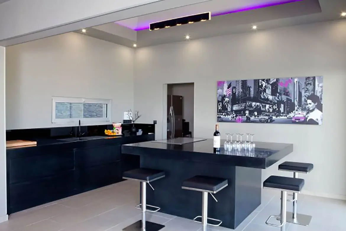 A modern minimalist kitchen features sleek black cabinetry, a large matching island with bar stools, recessed lighting with purple LED accent, and contemporary wall art.