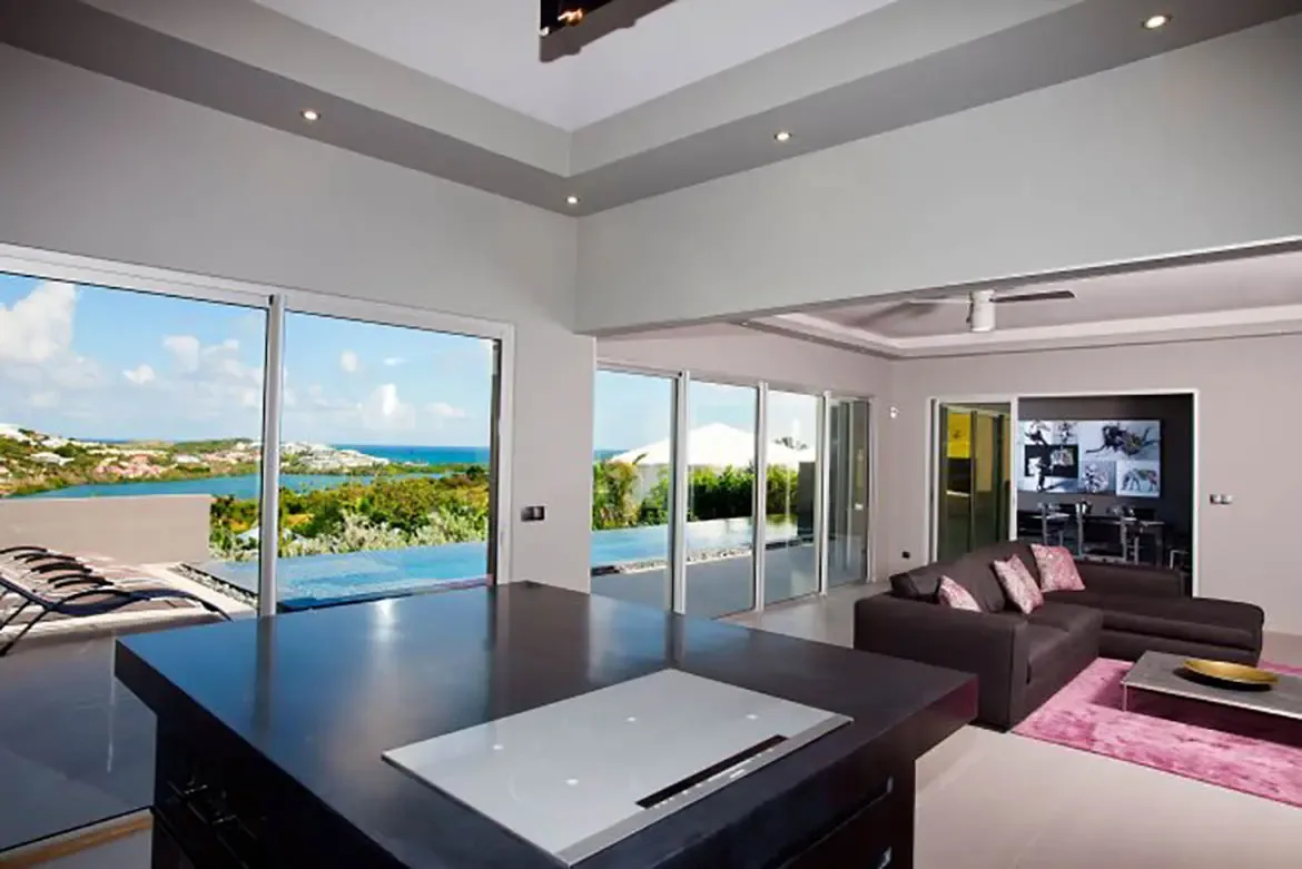 The open-plan living space features a modern kitchen island with induction cooktop, seating area with pink accent rug, and floor-to-ceiling glass doors revealing infinity pool and ocean view.