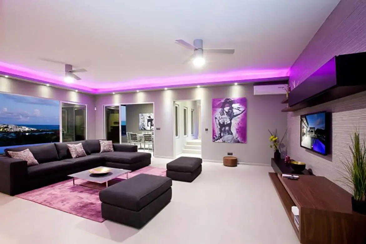 The modern living room features a large dark gray sectional, purple area rug, striking purple LED cove lighting, two ottomans, wall-mounted TV, and ocean view through expansive windows.