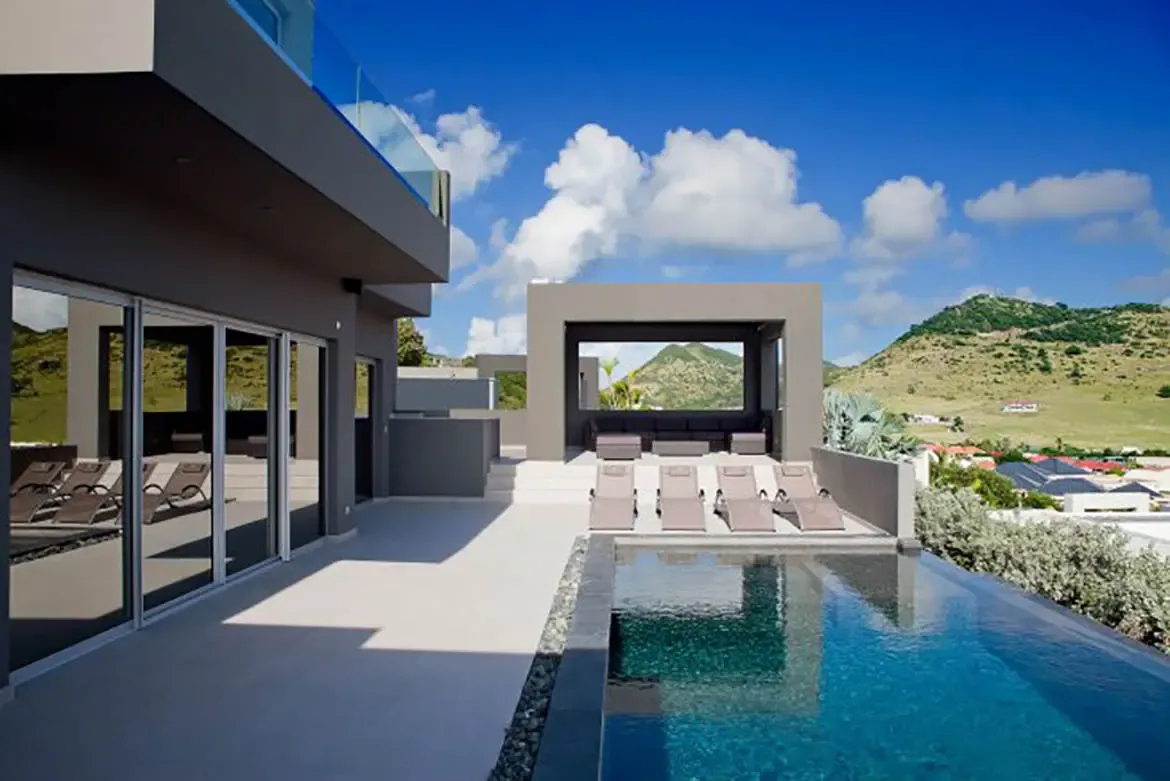The modern infinity pool area features sun loungers and a covered outdoor pavilion with seating, all set against stunning mountain views.