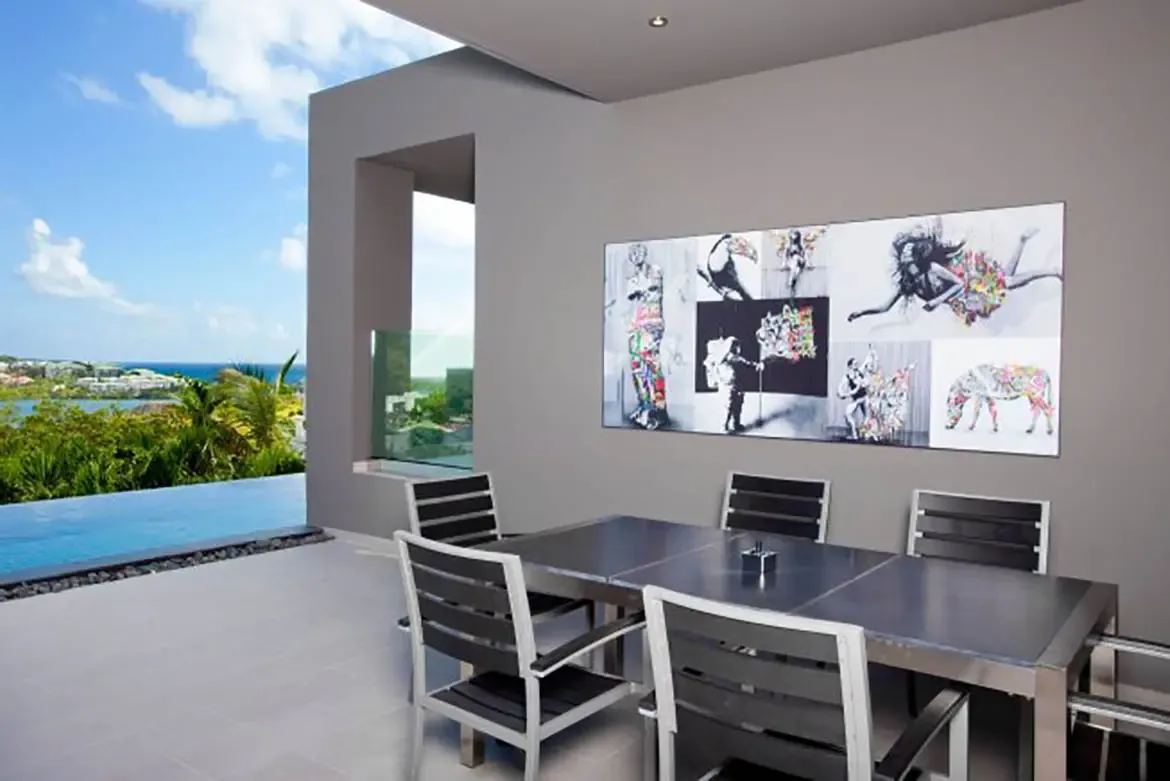A modern outdoor dining area with large collage artwork overlooks the infinity pool and scenic coastal view.