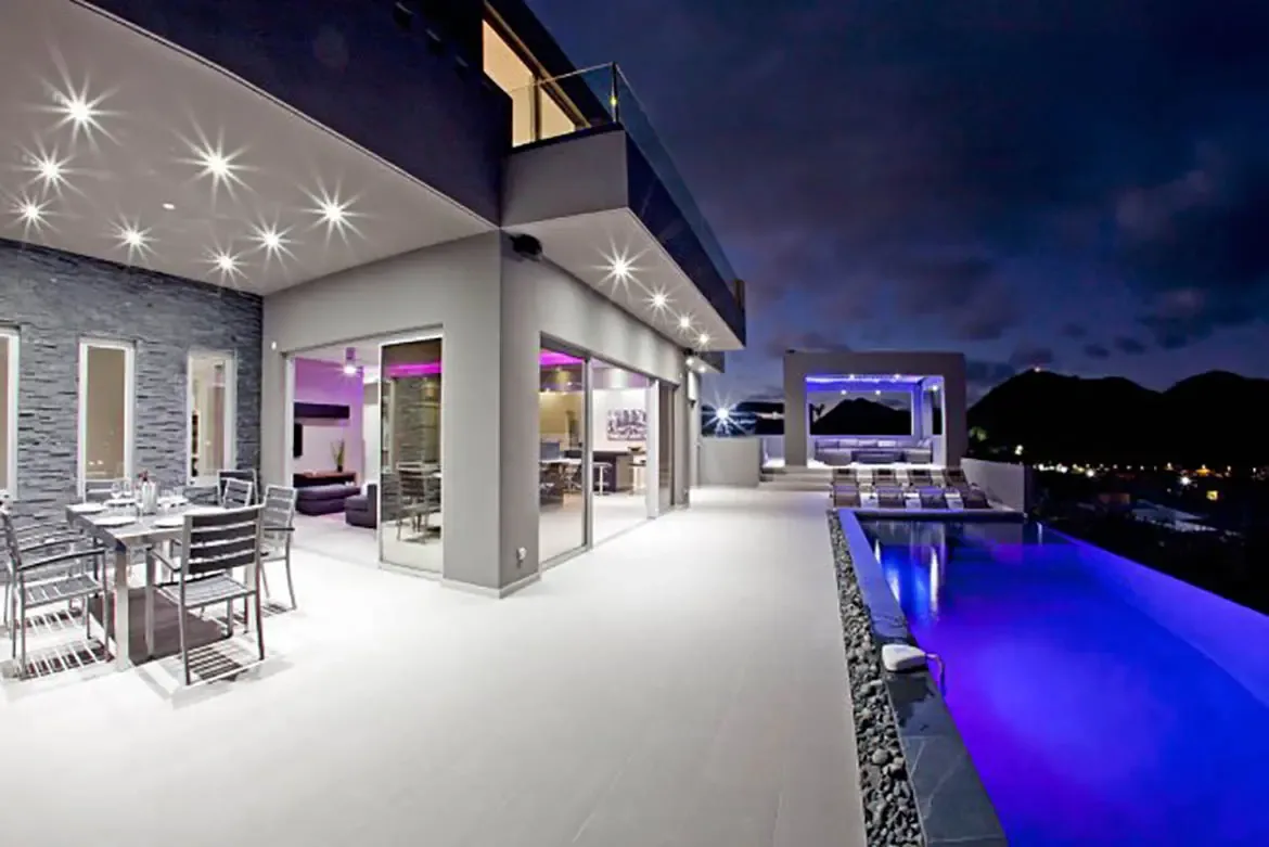 A luxurious night shot features an infinity pool with purple lighting, modern outdoor dining area, and sleek architectural lines against distant mountains backdrop.