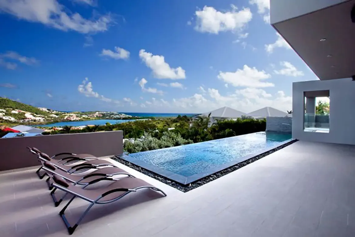 An elevated infinity pool overlooks a stunning coastal landscape and bay, with modern lounge chairs and contemporary villa architecture framing the view.