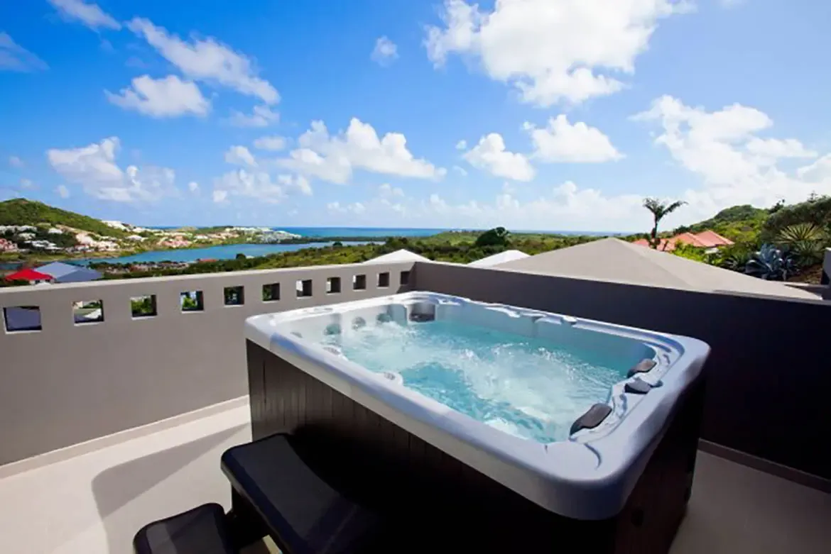 An outdoor terrace features a modern hot tub with steps, set against expansive view of tropical landscape, lagoon, and ocean under bright blue sky.