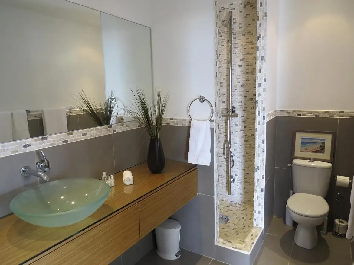 A modern bathroom featuring a floating wooden vanity with frosted glass vessel sink, large wall mirror, walk-in shower with mosaic tiles, and grey wall tiling with decorative borders.