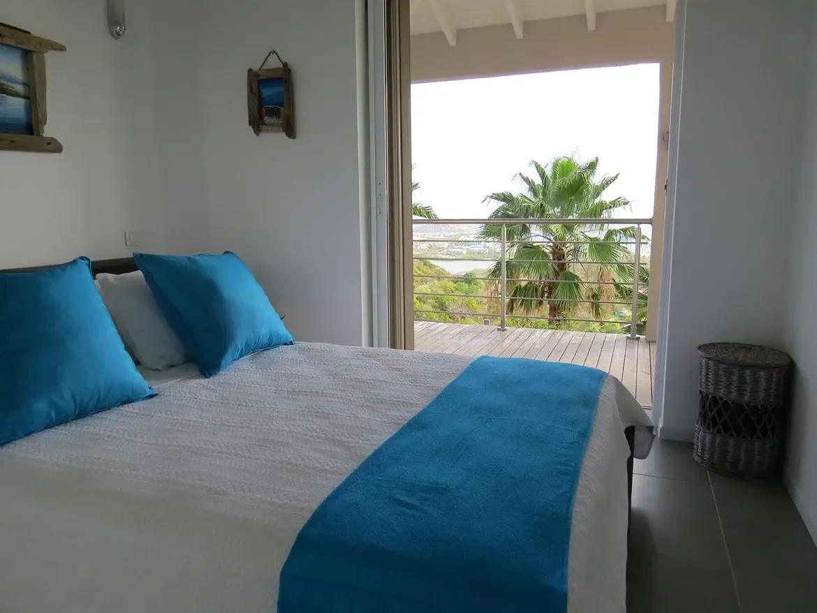 A bright bedroom featuring a white bed with turquoise accent pillows, sliding glass door opening onto a wooden deck overlooking lush tropical landscape with palm tree.
