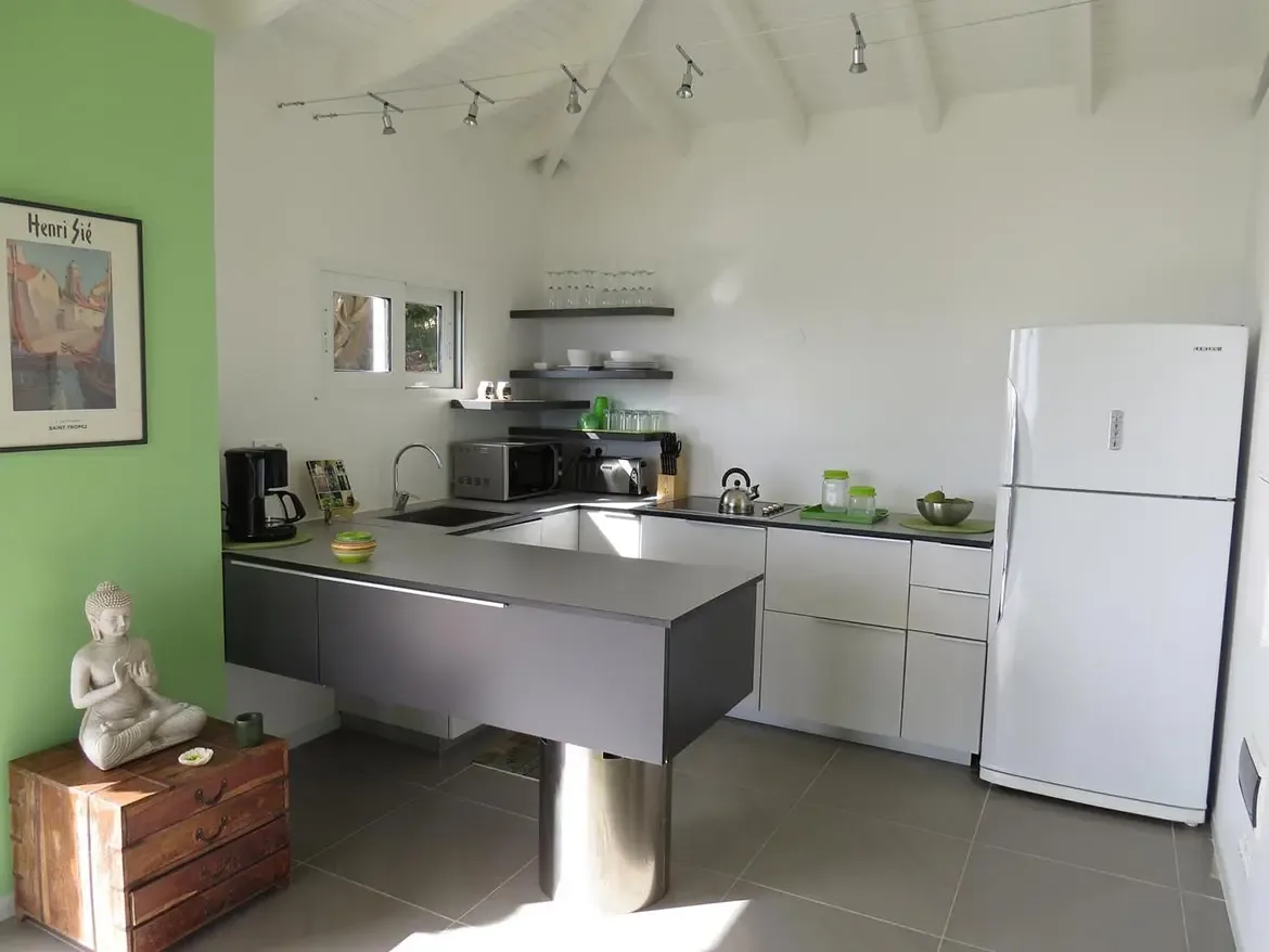 A modern L-shaped kitchen with white cabinetry, grey countertops, matching grey island bar, and a vibrant green accent wall with framed poster and Buddha statue.
