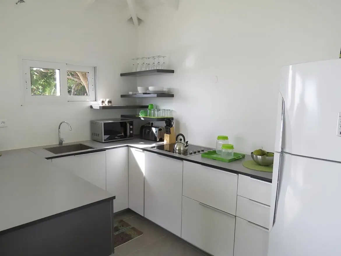 A modern L-shaped kitchen featuring white cabinetry, gray countertops, open wooden shelving, stainless steel sink, microwave, toaster, and stovetop with kettle.
