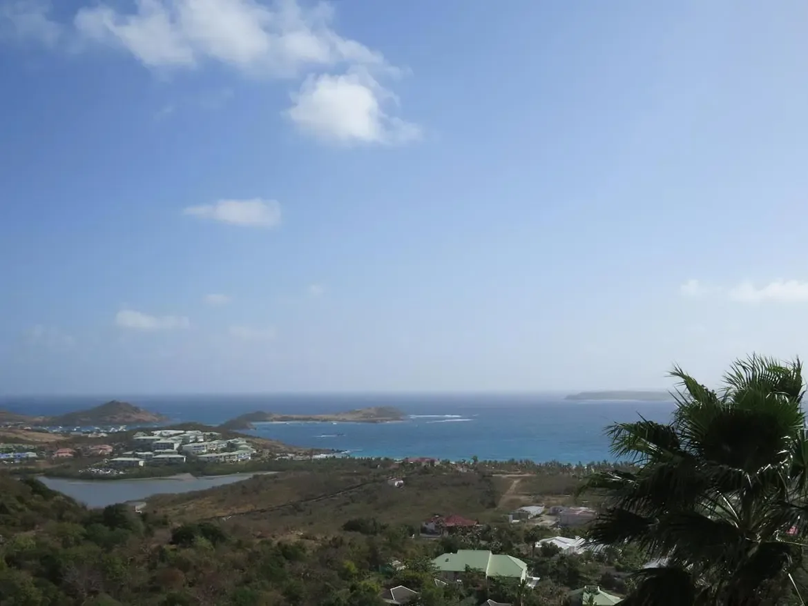 A wide-angle landscape shot overlooking a tropical bay with lush green hills, residential settlements, and distant islands under a bright blue sky.