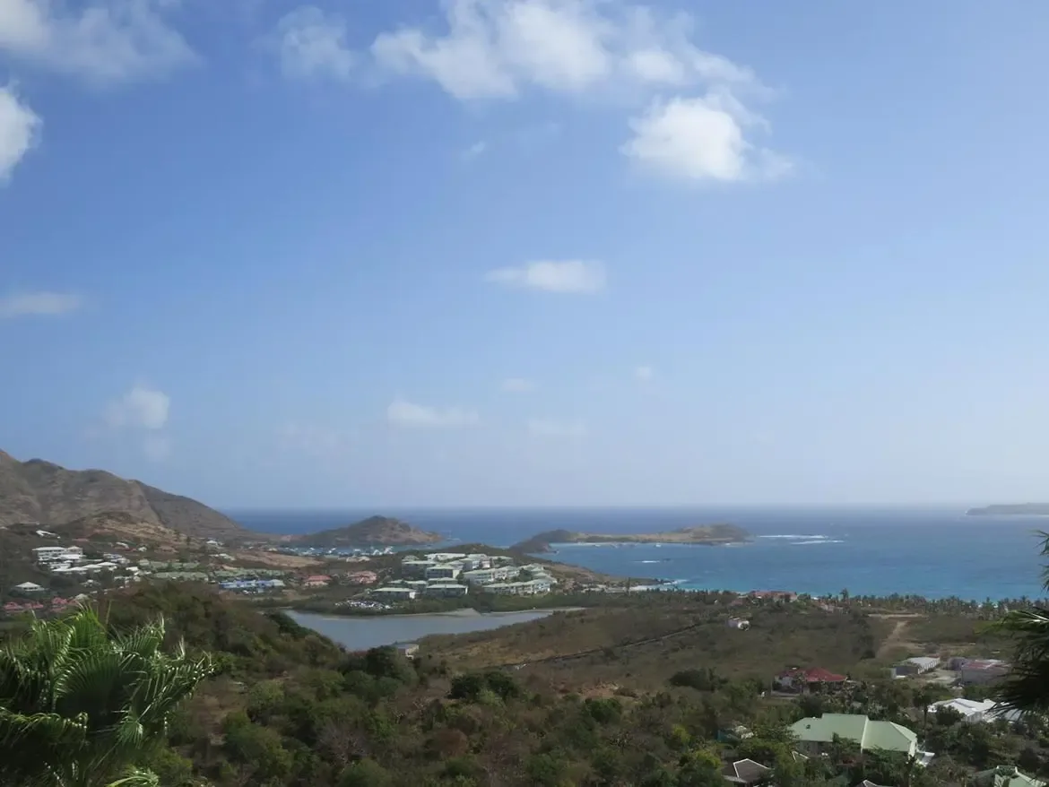 An elevated panoramic landscape view overlooking a coastal village, small inland lagoon, and the ocean under a bright partly cloudy sky.