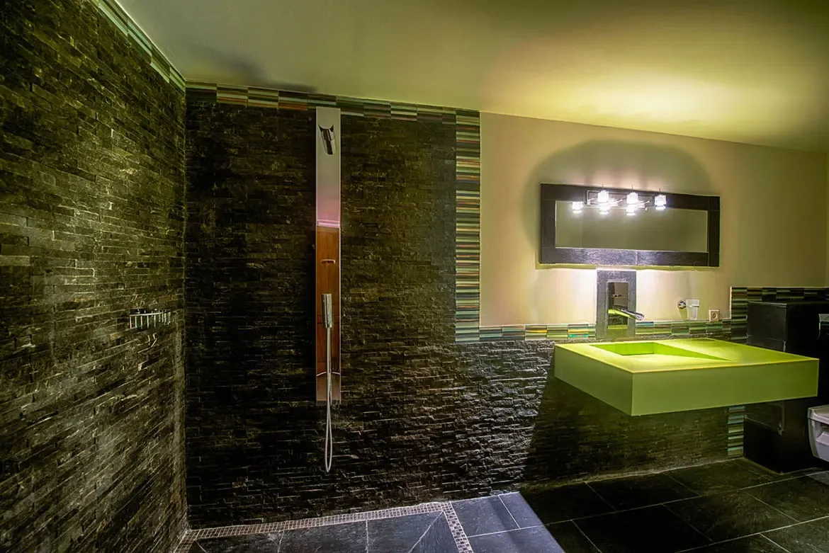 A contemporary bathroom with dark stacked-stone walls, walk-in shower, modern shower column, and lime-green floating sink with globe lighting.