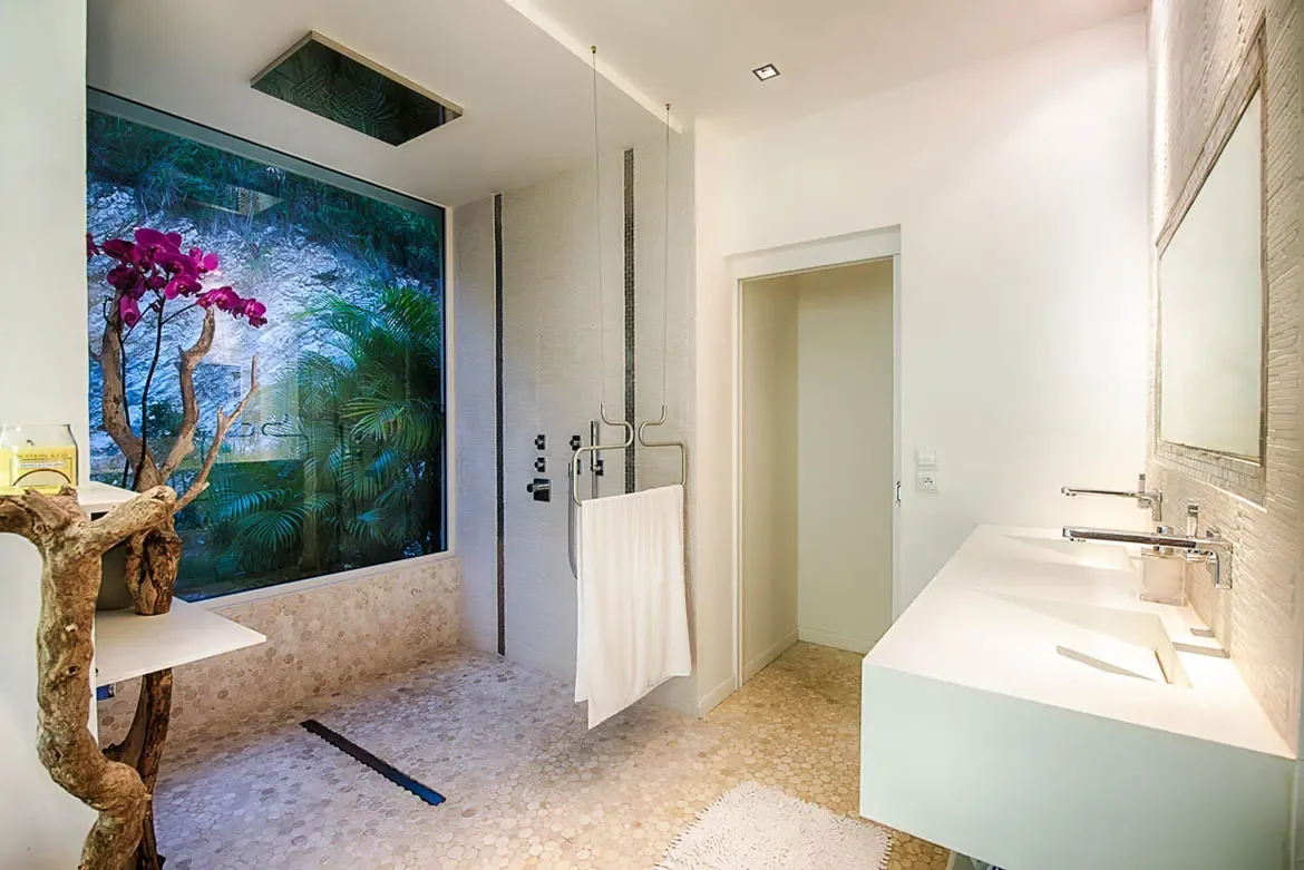 A spacious luxury bathroom featuring walk-in shower with pebble-stone flooring, ceiling rain shower, white double vanity, and floor-to-ceiling window with tropical views.