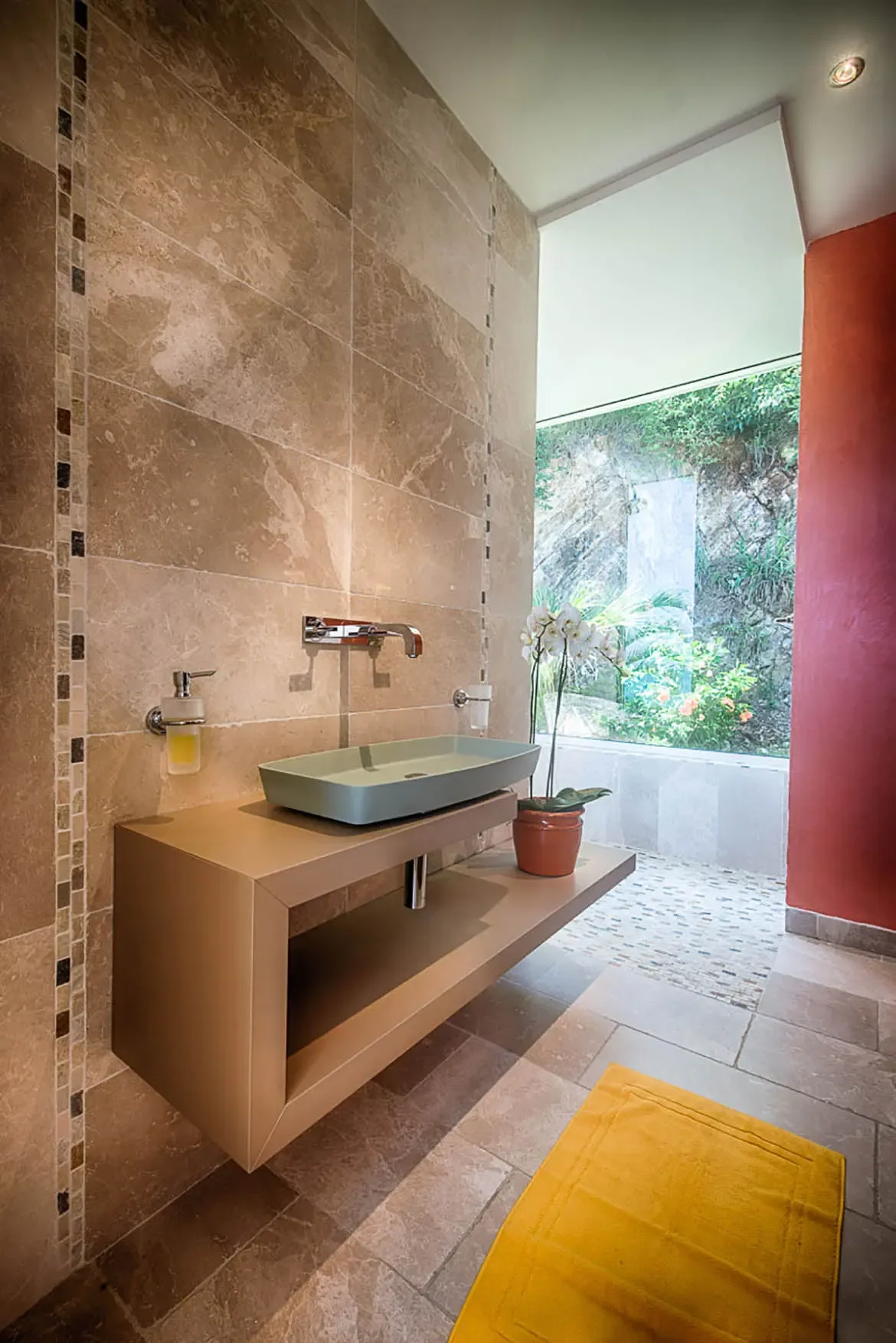 A modern bathroom featuring a floating gold-toned vanity with light green vessel sink, wall-mounted faucet, and large window overlooking lush outdoor greenery.