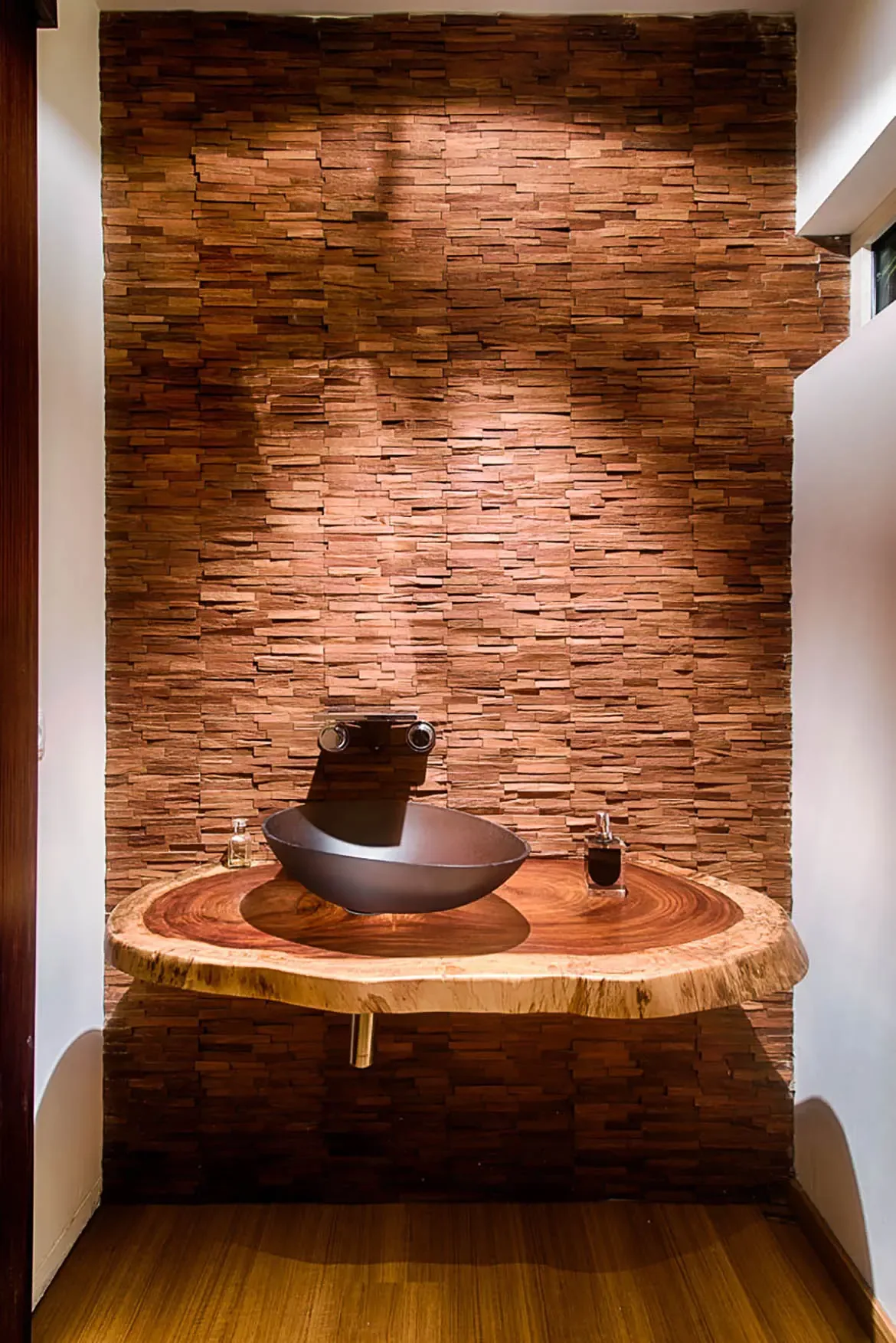 A closeup of floating live-edge wood vanity with dark boat-shaped vessel sink and wall-mounted faucets against textured stacked-stone accent wall.