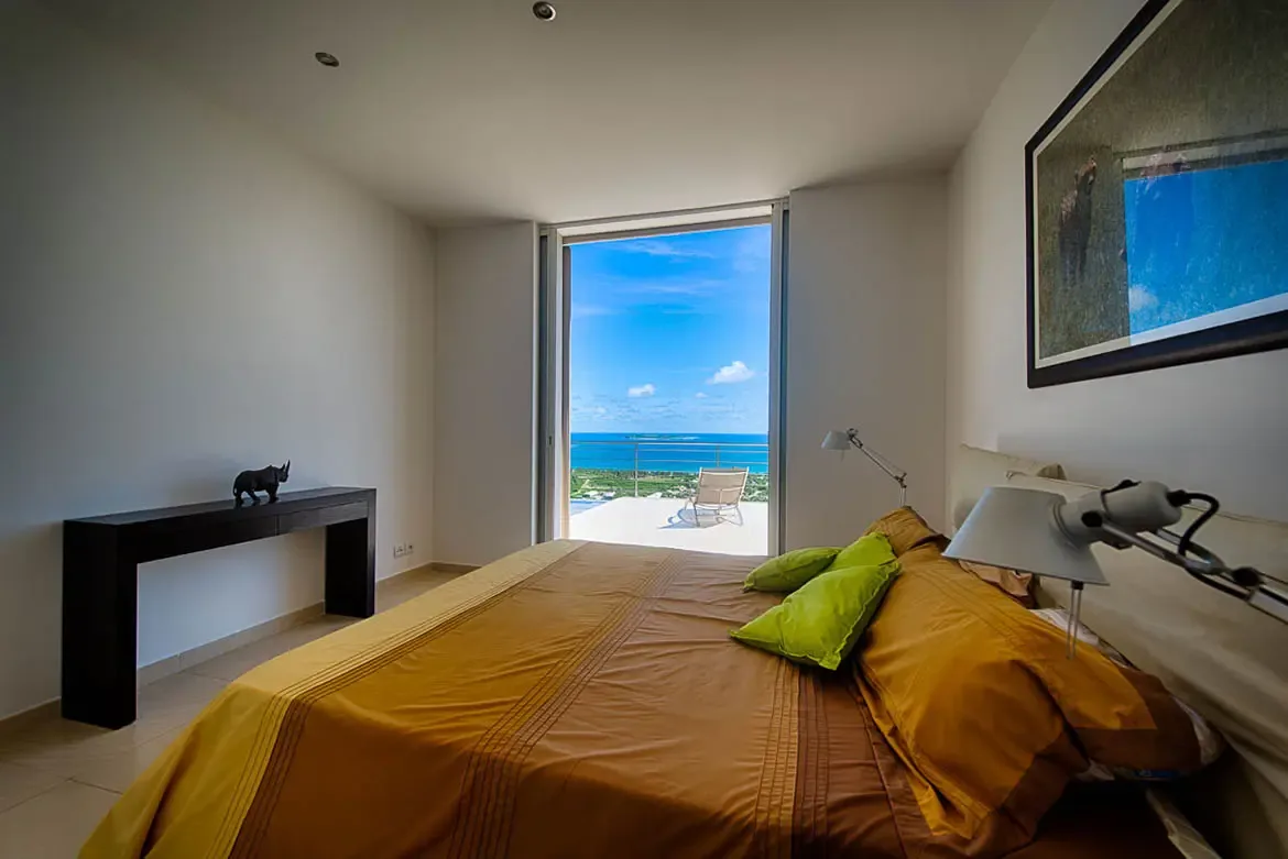 A modern bedroom with mustard-yellow bedding and green accent pillows, floor-to-ceiling opening to terrace with stunning panoramic turquoise ocean view.
