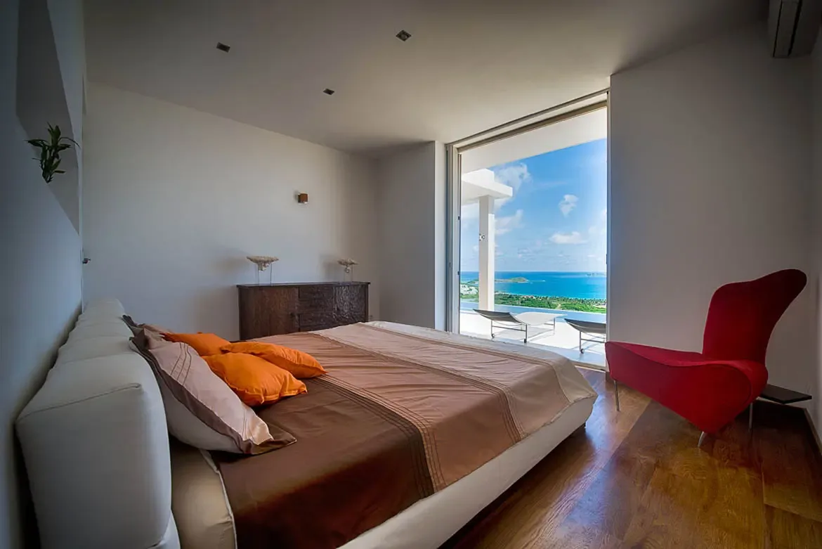 A modern minimalist bedroom with large bed, red designer armchair, opening directly onto a private terrace with panoramic ocean and coastline views.
